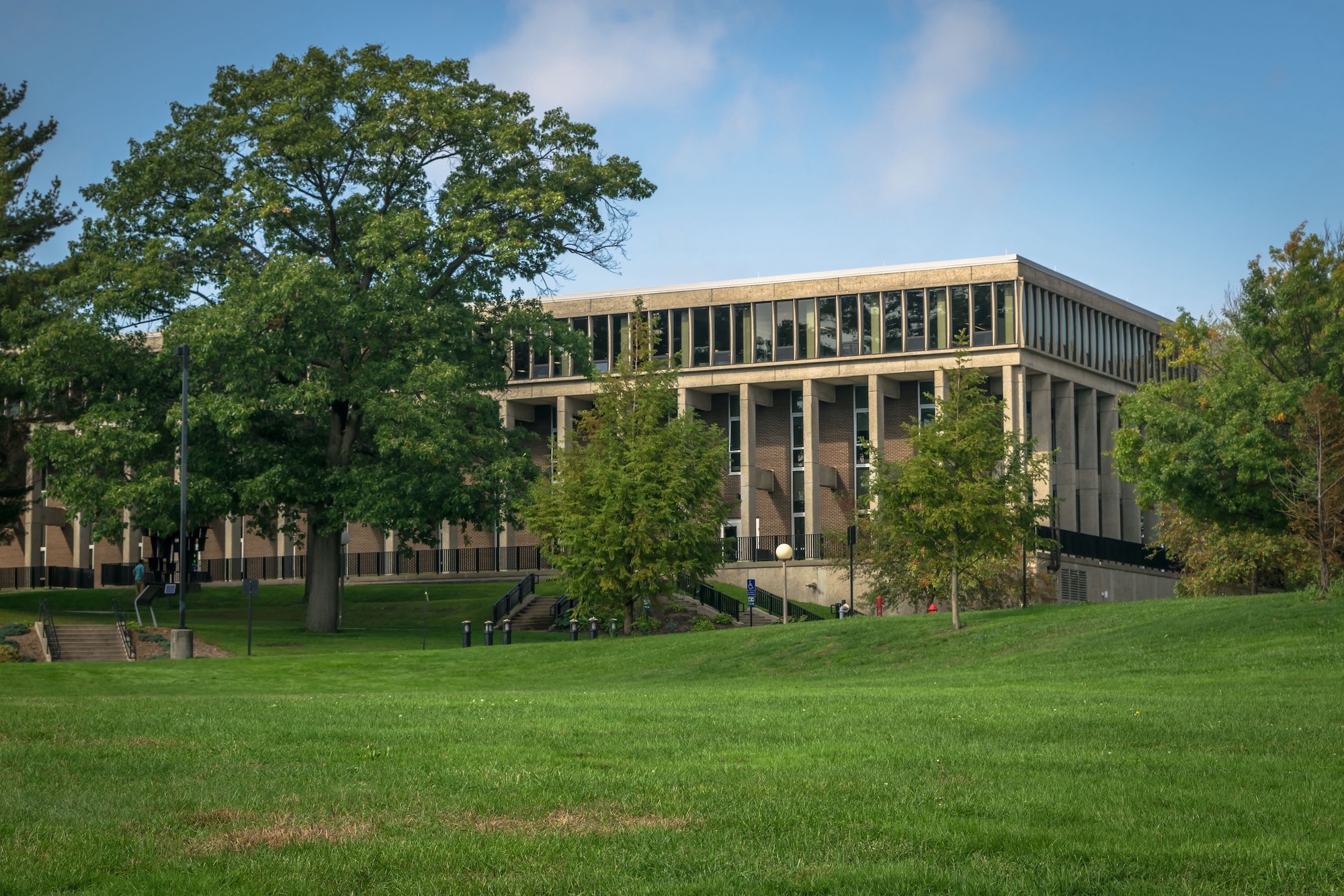 Large modern concrete campus building surrounded by green trees and a grassy lawn under a blue sky