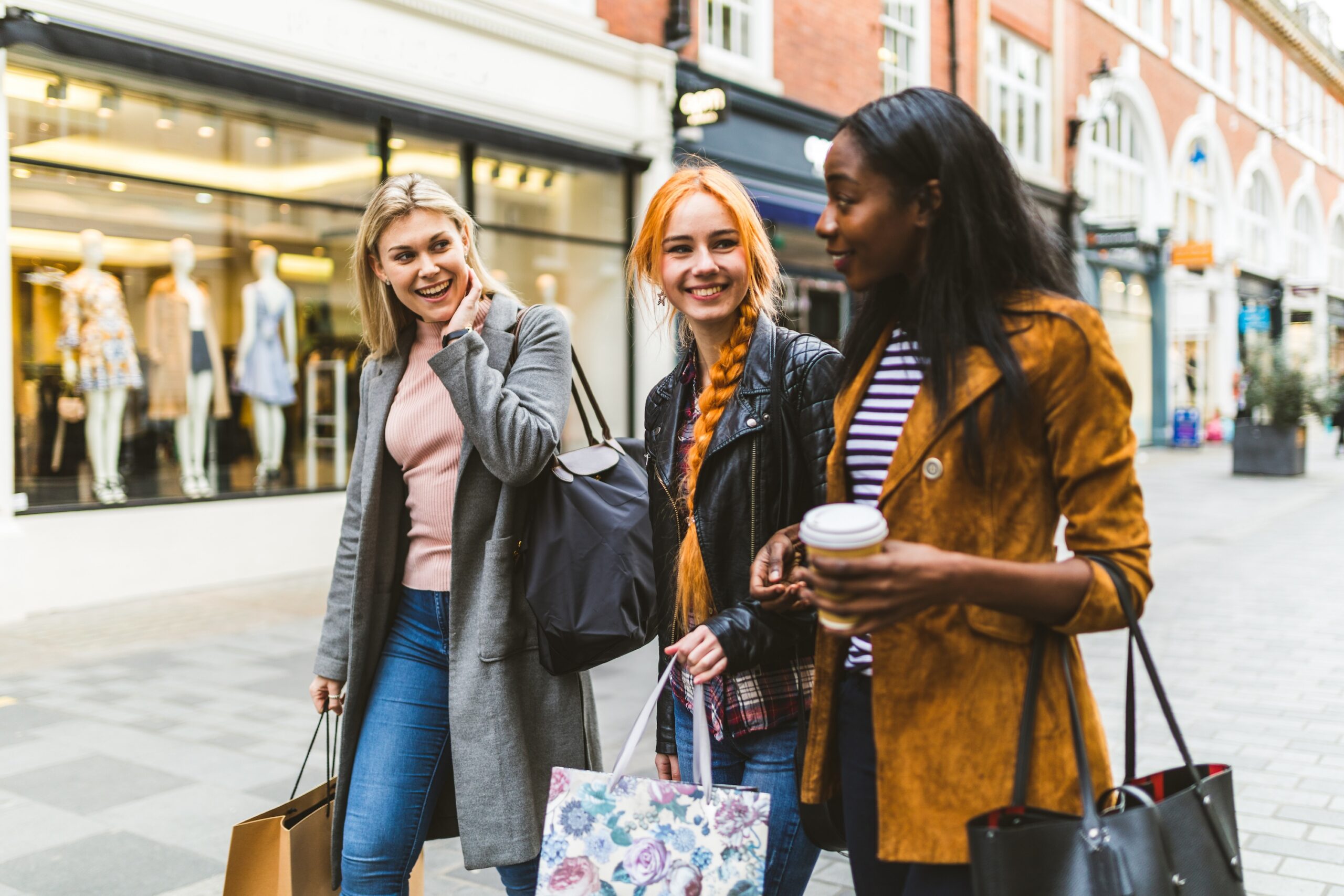 Three happy young women walking together on an outdoor shopping street holding shopping bags and coffee scaled